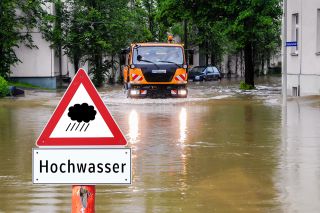 figure_photo: Hochwasserschild vor überfluteter Straße, Straßenmeisterfahrzeug im Hintergrund